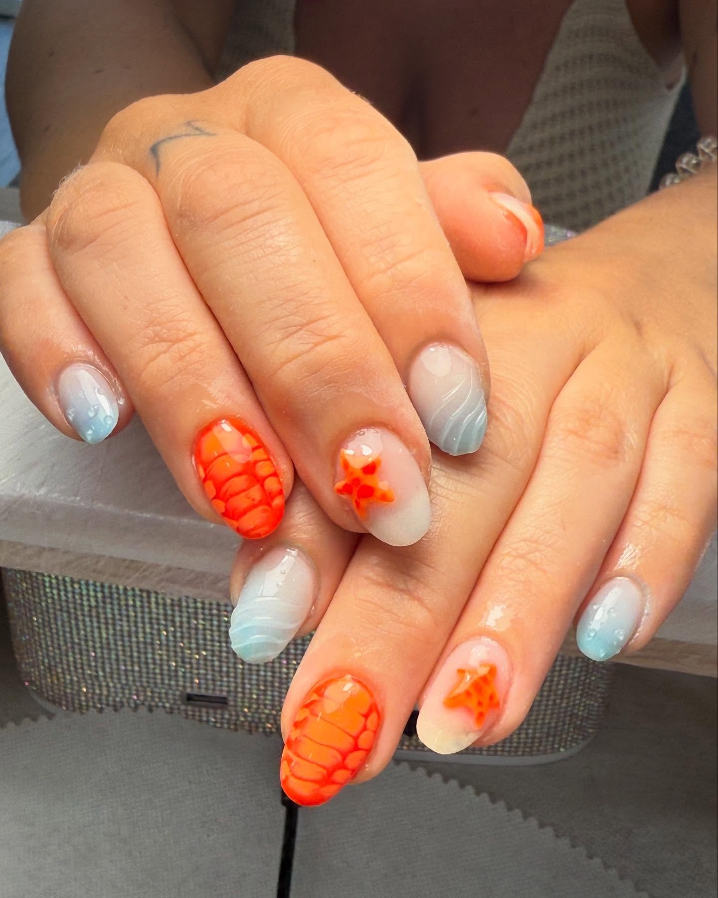 Manos con uñas decoradas en colores pastel azul y naranja. Algunas uñas tienen diseño de estrellas naranjas y patrón de escamas, otras están pintadas en degradado azul.